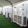 The image shows a row of large white HVAC/climate control units from United Rentals positioned along the interior wall of a white event tent. The units are lined up on a black mat surface beside artificial green turf flooring. Multiple electrical cables and connections are visible running along the base of the equipment.