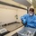 AFMES Joint MWD Lab ensures DoD kennels are meeting the standard A laboratory technician wearing full PPE (personal protective equipment) is working at a fume hood, pouring liquid from a dark bottle into a gray plastic bin containing metal canisters. Numerous small metal tins with perforated lids are arranged in rows on the fume hood surface, alongside a precision analytical balance. The setting appears to be a professional research or testing laboratory.