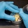 AFMES Joint MWD Lab ensures DoD kennels are meeting the standard A technician wearing blue nitrile gloves is handling a small metallic cylindrical component containing a gold/yellow-colored internal part, likely an electronic or mechanical assembly. A handheld tool with a blue indicator light is positioned nearby, suggesting a precision assembly or inspection task. The work is being performed on a dark laboratory or workbench surface.
