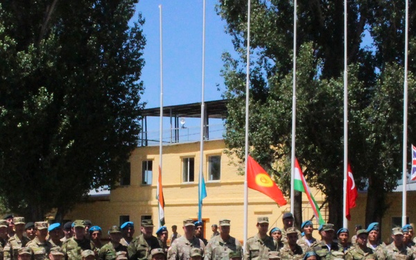 A large multinational military group photo taken outdoors at what appears to be a military installation or training facility. Personnel in various camouflage uniforms and blue berets are posed in formal rows, with several national flags visible in the background. The presence of multiple flags suggests a joint military exercise or international cooperation event.