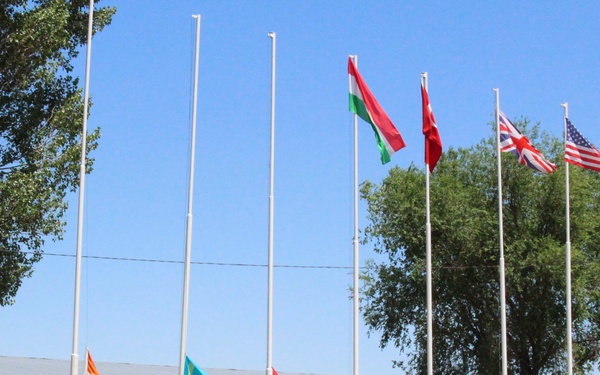 A military flag-raising ceremony is taking place at what appears to be a multinational military installation or training facility. Several soldiers in camouflage uniforms and blue berets are standing at salute beside flagpoles, raising the flags of multiple nations. The setting includes a yellow brick building in the background surrounded by trees under a clear blue sky.