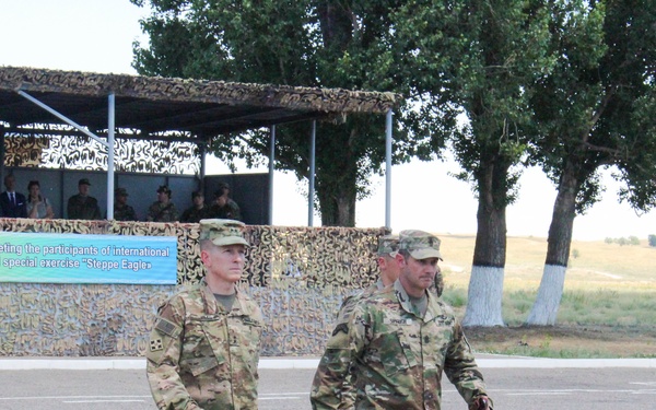 Two U.S. Army soldiers in MultiCam (OCP) camouflage uniforms are walking across a paved parade ground or assembly area. In the background, a covered viewing stand is occupied by military and civilian observers. A banner visible in the background references the international military exercise 'Steppe Eagle,' suggesting this is taking place in Central Asia, likely Kazakhstan.