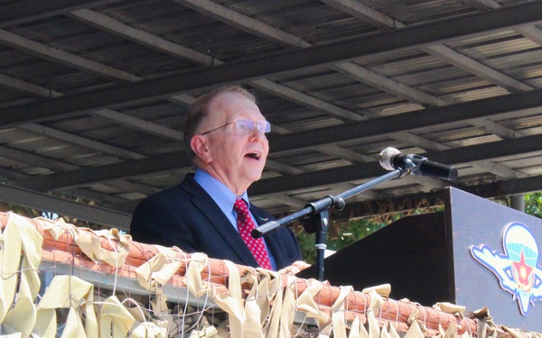 An elderly man in a dark suit and red tie is delivering a speech at a podium equipped with a microphone. The podium bears a military parachutist insignia, and the stage front is decorated with camouflage netting. A banner below the stage displays text in what appears to be Kazakh language, along with logos suggesting a US-Kazakhstan joint military exercise.