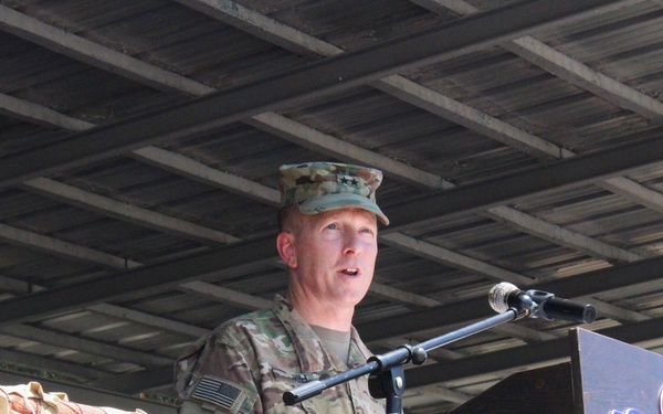 A U.S. Army officer in military combat uniform (OCP/MultiCam) is delivering a speech at a podium equipped with a microphone. The podium displays an airborne unit insignia, suggesting this is an official military ceremony or event. The elevated platform is decorated with camouflage netting along its railing.