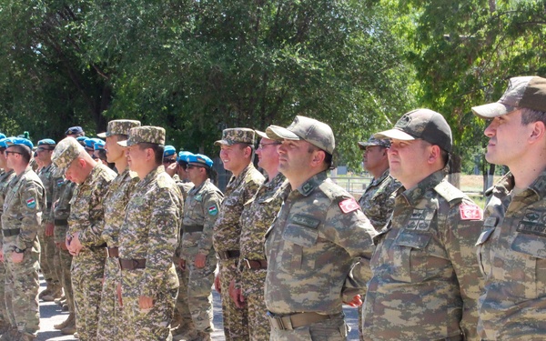 A formation of military personnel from multiple nations standing in rows on a paved surface, likely during a joint military exercise or ceremony. Soldiers are wearing different camouflage uniforms and headgear, with at least Turkish military personnel identifiable by flag patches on their sleeves. The setting appears to be an outdoor military installation with trees in the background.