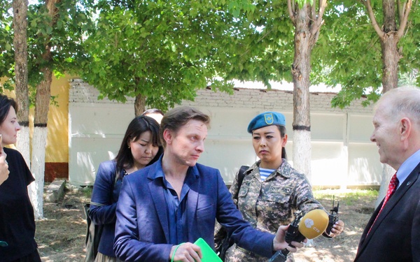 An outdoor press interview or media event is taking place in what appears to be a military or institutional compound. A civilian journalist holding a green notepad is conducting an interview with a man in a dark suit and red tie, while a female soldier in camouflage uniform and blue beret holds a microphone. A camera crew and additional observers are present in the background.