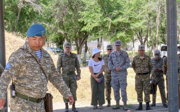 A military officer wearing a blue beret and camouflage uniform appears to be conducting a briefing or demonstration over a sand table model, which is used for tactical planning and instruction. A group of observers including military personnel in various uniforms and a few civilians stand watching in the background. The setting appears to be an outdoor military training or exercise area.