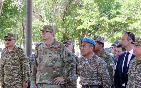 A group of military personnel and civilians are gathered outdoors, apparently observing a briefing or demonstration. The group includes soldiers in multiple different camouflage uniforms suggesting a multinational military exercise or meeting. Small white markers labeled 'UN' are visible on the ground in the foreground.