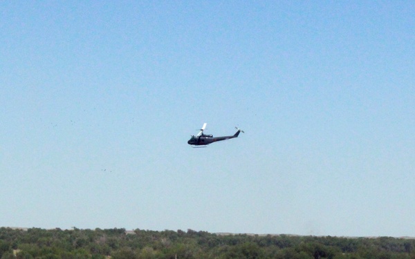 A military helicopter flies over what appears to be a mock village or training compound consisting of brightly colored small structures with multicolored roofs. The scene includes military vehicles positioned around the perimeter of the compound, set against a backdrop of trees and open terrain. This appears to be a military training exercise or demonstration area.
