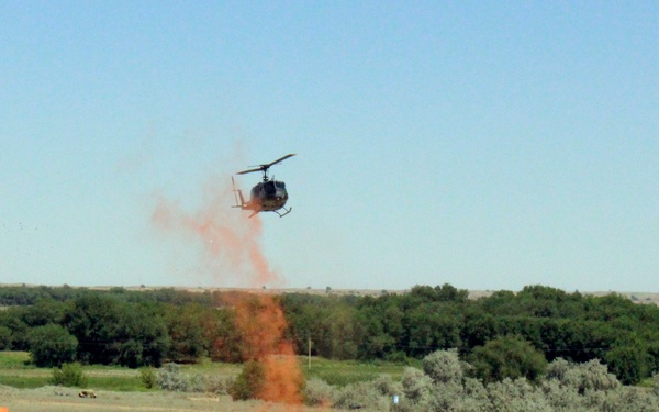 A military exercise or demonstration is underway in an open field, featuring a helicopter hovering above while orange smoke grenades or flares mark positions on the ground below. Ground vehicles, likely military utility trucks or HMMWVs, are positioned in the field surrounded by the orange smoke. The scene includes a mock village or training area with colorful small structures on the left and blue tents on the right, suggesting an organized training event.