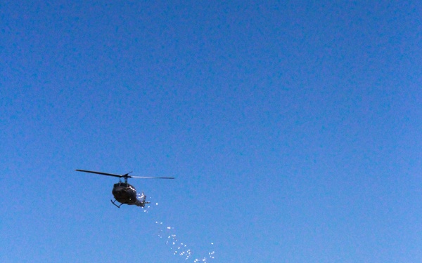 A dark-colored helicopter is captured in flight against a clear blue sky, dispersing a trail of small white objects that fall in a curved arc below it. The helicopter appears to be at moderate altitude above a treeline and what appears to be a small settlement or town visible at the bottom of the image. The dispersal trail suggests the helicopter is actively releasing items, possibly leaflets, seeds, or similar lightweight materials.