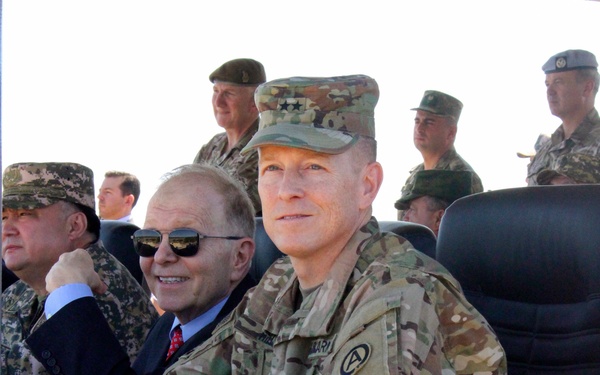 A group of military personnel and at least one civilian are seated and standing at what appears to be an official military observation or briefing event. The foreground features a US Army officer in MultiCam/OCP uniform with two-star (Major General) rank insignia on his cap and an 'A' (US Army Central/Third Army) shoulder patch. Multiple nationalities of military uniforms are visible, suggesting a multinational military gathering or exercise observation.