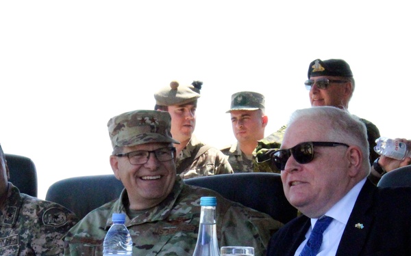 A formal military and diplomatic gathering showing officials seated at a conference table, likely during a multinational military exercise or official ceremony. Three primary figures are seated in the foreground, including military personnel and a civilian dignitary, with name placards visible. Additional military personnel from multiple nations are visible in the background.