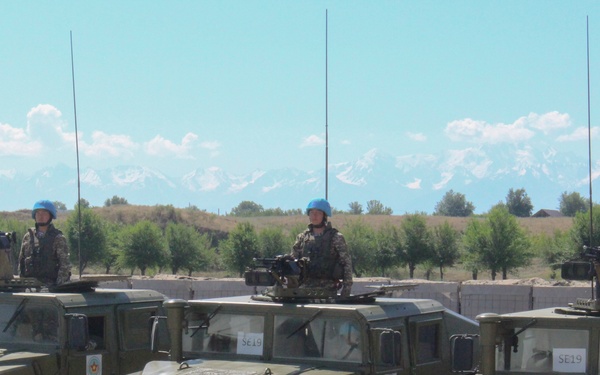 Three military HMMWV (Humvee) vehicles are lined up in a formation on a gravel surface, each with a soldier standing in the turret position wearing blue UN-style helmets. The vehicles bear license plates reading '7287 KA' and '7288 KA' and display 'SE19' markings on their windshields. Snow-capped mountains are visible in the background, suggesting a Central Asian location.