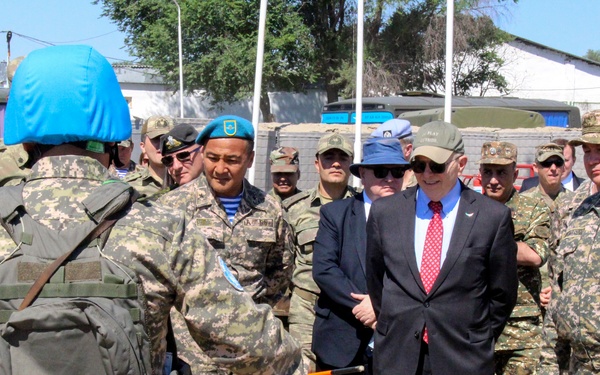 A group of military personnel in camouflage uniforms, along with a civilian in a dark suit and red tie, are gathered around a table with a scale model or display. The scene appears to be an outdoor military briefing or demonstration at a military installation. Multiple nationalities appear to be represented, with some personnel wearing blue UN-style berets.