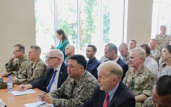 A formal military and civilian meeting or briefing is taking place in a conference room. Participants include U.S. Army personnel in camouflage uniforms and civilians in business attire, seated at a long table with name placards. The setting appears to be a multinational or bilateral military cooperation event, likely in Central Asia based on visible Cyrillic name placards and mixed uniform styles.