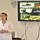 U.S. Transportation Command holds training on TMS prototype at Fort McCoy A woman is presenting in a meeting or briefing room, standing beside a large Pioneer flat-screen television displaying a U.S. Army Logistics PowerPoint slide. The presentation slide is titled 'Data Capture TMS Prototype, Ft McCoy' and includes military imagery such as a Black Hawk helicopter, soldiers, and armored vehicles. A whiteboard with partial handwritten notes is visible in the background.