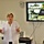 U.S. Transportation Command holds training on TMS prototype at Fort McCoy A woman is presenting at what appears to be a U.S. Army Logistics briefing in a conference or training room. The presentation slide on the Pioneer flat-screen TV behind her reads 'Data Capture TMS Prototype, Ft McCoy' under the U.S. Army Logistics banner. She is gesturing with both hands while speaking, with a whiteboard visible in the background.