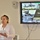 A woman is presenting at a briefing or meeting, holding papers and standing beside a large display screen. The screen shows a U.S. Army Logistics presentation slide titled 'Data Capture TMS Prototype, Ft Mc[Coy]' dated 3-7 June 2019. Behind her, a whiteboard is partially visible with numbered items written in red marker.