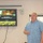 U.S. Transportation Command holds training on TMS prototype at Fort McCoy A middle-aged man in a blue plaid shirt is presenting at what appears to be a U.S. Army logistics briefing. The presentation slide on the wall-mounted TV displays 'Data Capture TMS Prototype, Ft McCoy' under a U.S. Army Logistics header. The setting is an indoor conference or briefing room with a whiteboard visible in the background.