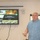 U.S. Transportation Command holds training on TMS prototype at Fort McCoy A middle-aged man in a blue plaid shirt is presenting at what appears to be a U.S. Army logistics briefing. The presentation slide on the TV monitor behind him reads 'Data Capture TMS Prototype, Ft McCoy' under the U.S. Army Logistics banner. The setting appears to be a conference or meeting room.