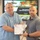 Two men are posing together while holding a certificate issued by the Transportation Management System (TMS) made out to 'David Eckland'. They are standing in front of a presentation screen displaying slides related to 'Data Capture TMS Prototype, Ft [location partially obscured]'. The setting appears to be a formal recognition or training completion ceremony.