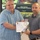U.S. Transportation Command holds training on TMS prototype at Fort McCoy Two men are jointly holding a Certificate of Recognition issued by the Transportation Management System (TMS) to an individual named David Eckord. The ceremony appears to be taking place in an office or conference room with a U.S. Army-branded presentation displayed on a screen in the background, referencing 'Data Capture TMS Prototype, Ft McCoy.' The certificate also features what appears to be a 'TMS Outstanding Contributor Award' medallion.