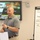 U.S. Transportation Command holds training on TMS prototype at Fort McCoy A man in a gray polo shirt is presenting in what appears to be a military logistics training or briefing session. Behind him is a monitor displaying a 'U.S. Army Logistics' slide with the text 'TMS McCoy,' and to his right is a whiteboard/flipchart with URLs and numbered items. He is holding a sheet of paper and appears to be speaking to an audience.