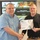 U.S. Transportation Command holds training on TMS prototype at Fort McCoy Two men are shaking hands while jointly holding a Certificate of Recognition issued by Transportation Management Systems to 'Douglas Altman.' The ceremony appears to be taking place in a conference or briefing room with a projected U.S. Army presentation visible in the background titled 'Data Capture TMS Prototype, Ft McCoy.' The certificate includes an Outstanding Contribution Award medallion seal.