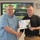 U.S. Transportation Command holds training on TMS prototype at Fort McCoy Two men are shaking hands while jointly holding a Transportation Management System (TMS) certificate awarded to 'Douglas Altman.' The ceremony appears to take place in a conference or briefing room with a U.S. Army presentation displayed on a screen behind them. The slide on the screen references 'Data Capture TMS Prototype, Ft McCoy' dated 5-7 June 2009.