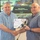 U.S. Transportation Command holds training on TMS prototype at Fort McCoy Two men are shaking hands while jointly holding a certificate from the Transportation Management System (TMS). The certificate is made out to 'Dean Muller' and appears to be an outstanding contribution award. The event appears to be related to a 'Data Capture TMS Prototype, Ft McCoy' presentation visible on the screen behind them.