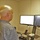 U.S. Transportation Command holds training on TMS prototype at Fort McCoy A bald man wearing a gray plaid short-sleeve polo shirt is standing at a sit-stand desk converter, working at a dual-monitor computer setup. He is using a mouse with his right hand while facing two Dell monitors mounted on a standing desk riser. The workspace appears to be a small office cubicle or private office environment.