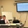 U.S. Transportation Command holds training on TMS prototype at Fort McCoy A woman is seated at a conference table working on a laptop in what appears to be a meeting or training room. A large wall-mounted Pioneer display screen shows what appears to be a spreadsheet or document with multiple columns. A decorated cake and conference phone are also visible on the table.