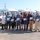 U.S. Transportation Command holds training on TMS prototype at Fort McCoy A group of approximately eleven adults are posed together in a parking lot in front of a row of white Ford Transit vans. Most individuals are holding certificates or plaques, suggesting a recognition or award ceremony. The setting appears to be a fleet vehicle lot or transportation facility.