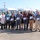 U.S. Transportation Command holds training on TMS prototype at Fort McCoy A group of approximately eleven adults pose for a photo in a parking lot in front of a row of white Ford Transit vans. Most individuals are holding certificates or plaques, suggesting a recognition or award ceremony. The setting appears to be a commercial or fleet vehicle lot on a clear, sunny day.