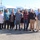 U.S. Transportation Command holds training on TMS prototype at Fort McCoy A group of approximately eleven adults poses for a group photo in a parking lot in front of a fleet of white Ford Transit vans. The setting appears to be a commercial or fleet vehicle lot, likely during a vehicle delivery or organizational event. The photo appears to be an official or commemorative group shot taken outdoors on a clear, sunny day.