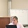 U.S. Transportation Command holds training on TMS prototype at Fort McCoy A woman in a pink blazer is seated at a table working on a black laptop while gesturing with her right hand, suggesting she is presenting or explaining something. Behind her is a whiteboard with a numbered diagram (1-5) written in red marker, indicating a training or meeting environment. The setting appears to be a conference or classroom room.