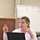 U.S. Transportation Command holds training on TMS prototype at Fort McCoy A woman is seated at a white table in what appears to be a classroom or training room, actively speaking and gesturing with both hands raised. She has a laptop computer open in front of her and a red wireless mouse to her left. A whiteboard with numbered sections and written content is visible in the background.
