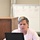 U.S. Transportation Command holds training on TMS prototype at Fort McCoy A middle-aged woman with short gray-blonde hair sits at a table working on a black laptop in what appears to be a meeting or training room. She is wearing a light pink cardigan over a white top with a gold necklace, and appears to be speaking or presenting. Behind her is a whiteboard with numbered sections containing handwritten red text and diagrams.