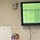 U.S. Transportation Command holds training on TMS prototype at Fort McCoy A woman in a light pink cardigan sits at a desk with a laptop in what appears to be a conference or training room. Behind her is a whiteboard with handwritten notes about freight logistics, and above is a wall-mounted monitor displaying a Microsoft Excel spreadsheet with green-highlighted cells related to capability assessments at Fort McCoy. The setting suggests a professional training or planning session.