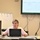 U.S. Transportation Command holds training on TMS prototype at Fort McCoy A woman is presenting in a conference or training room, seated at a table with a laptop and mouse. Behind her, a large wall-mounted Pioneer display shows a spreadsheet or assessment matrix document. A whiteboard to her left contains a numbered process flow diagram with handwritten labels.