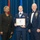 Airman Leadership School distinguished graduate A U.S. Air Force Airman is being recognized at what appears to be an Airman Leadership School graduation ceremony. The center individual holds a framed diploma from the United States Air Force Airman Leadership School, flanked by a civilian woman in a black suit and a senior non-commissioned officer in dress blues. The setting features an American flag and blue ceremonial draping in the background.