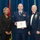 Airman Leadership School distinguished graduate A formal military graduation ceremony at the United States Air Force Noncommissioned Officer Leadership School. A Senior Airman identified on the diploma as 'Joshua R. Alley' holds a framed diploma while posing with two other individuals, one in civilian attire and one in a senior NCO dress uniform. The setting features multiple flags and a blue draped backdrop consistent with an official military ceremony.