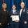 Airman Leadership School distinguished graduate A United States Air Force graduation ceremony at the Airman Leadership School, where a female airman is being recognized with a diploma. She is flanked by two presenters — a civilian woman in a black suit on the left and a senior enlisted male officer in dress uniform on the right. The recipient is holding a framed diploma and wearing a medal/lanyard around her neck.