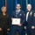 Airman Leadership School distinguished graduate A formal military graduation or awards ceremony featuring three individuals posing for a photo. The central figure, a U.S. Air Force enlisted member wearing a medal and ribbon, holds a framed diploma from the United States Air Force Airmen Leadership School. He is flanked by a civilian woman in a black blazer on the left and a senior U.S. Air Force Chief Master Sergeant in dress uniform on the right.