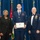Airman Leadership School distinguished graduate Three individuals pose for a formal photograph at what appears to be a United States Air Force ceremony. The central figure, a uniformed airman, holds a framed diploma or certificate and wears a medal on a ribbon around his neck. He is flanked by a civilian woman in black attire on his left and a senior non-commissioned officer in dress uniform with multiple medals on his right.