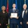 Airman Leadership School distinguished graduate A formal military ceremony showing three individuals posing for a photo. A young airman in the center holds a framed United States Air Force Airman Leadership School diploma, flanked by a civilian woman in black attire on the left and a senior non-commissioned officer in dress uniform on the right. The setting features a blue velvet backdrop with multiple flags including the US flag and what appears to be the Jordanian flag.