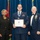 Airman Leadership School distinguished graduate A formal military graduation or awards ceremony featuring three individuals posing for a photo. The central figure, a uniformed Air Force member wearing a medal and holding a framed diploma, is flanked by a civilian woman in a black suit and a senior enlisted Air Force member in dress uniform. The diploma reads 'United States Air Force Airman Leadership School' and is made out to 'SrA Benjamin B. Ockert'.