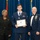 Airman Leadership School distinguished graduate A military graduation or awards ceremony featuring three individuals posing for a formal photograph. The central figure, a young airman in dress blues, is holding a framed diploma and wearing a medal around his neck. He is flanked by a woman in civilian attire and a senior non-commissioned officer in dress uniform with multiple medals and ribbons.