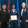 Airman Leadership School distinguished graduate Three individuals pose for a formal photograph at what appears to be a United States Air Force Airman Leadership School graduation ceremony. The central figure, a female airman in dress uniform, holds a framed diploma and wears a medal around her neck. She is flanked by a civilian woman in a black suit on the left and a senior enlisted Air Force officer in dress uniform on the right.